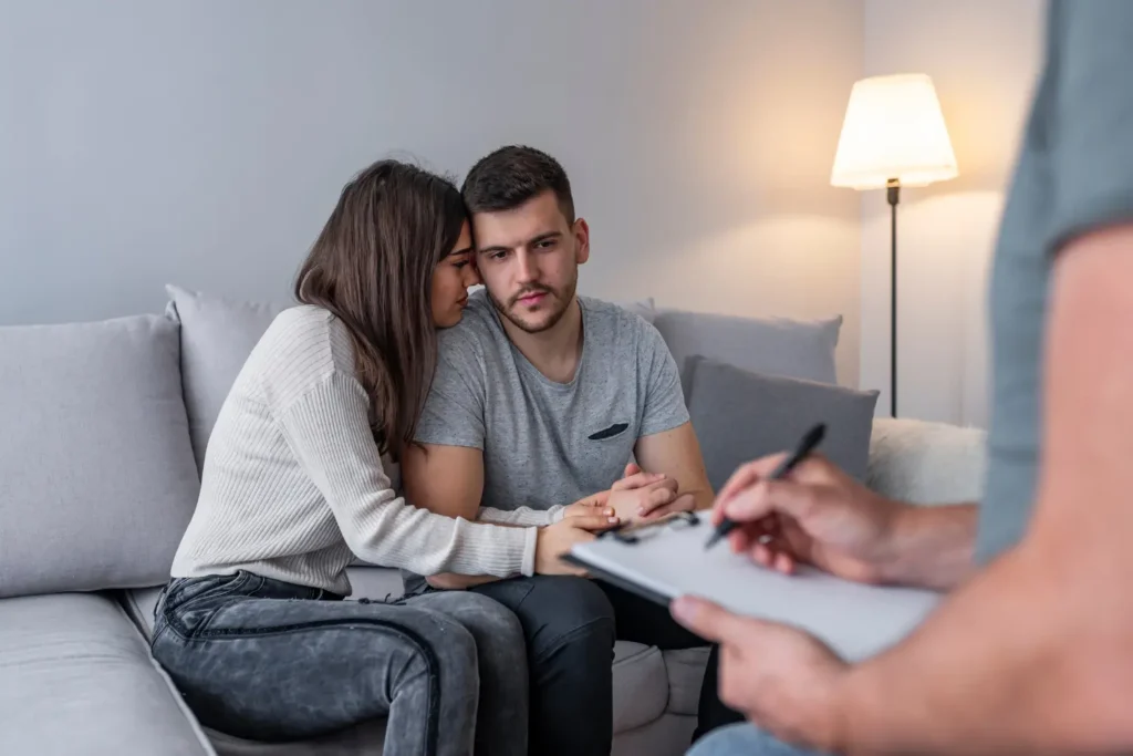 a wife sitting on a couch hugging and supporting her husband in therapy