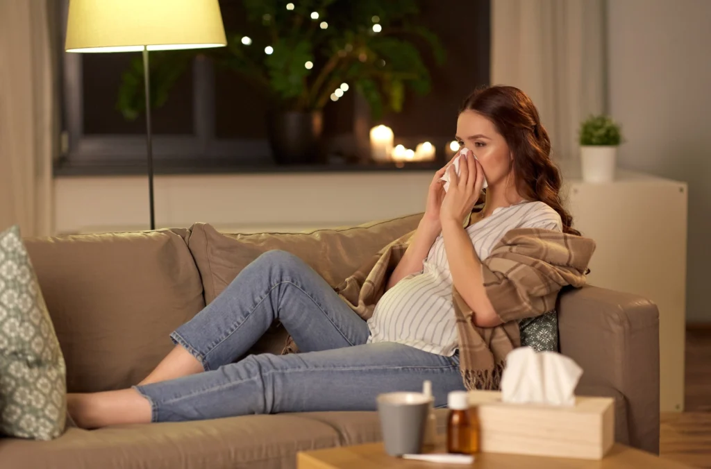 Dextromethorphan During Pregnancy: A pregnant woman on a couch blowing her nose with medication on the table in front of her.
